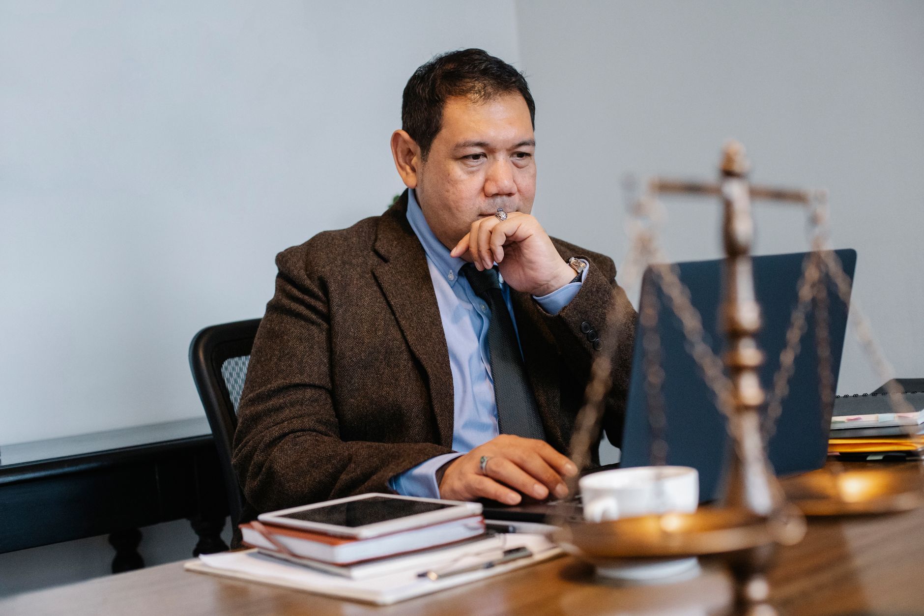 Serious adult Asian male judge in formal clothes browsing netbook and touching chin in thoughts while working in modern law office