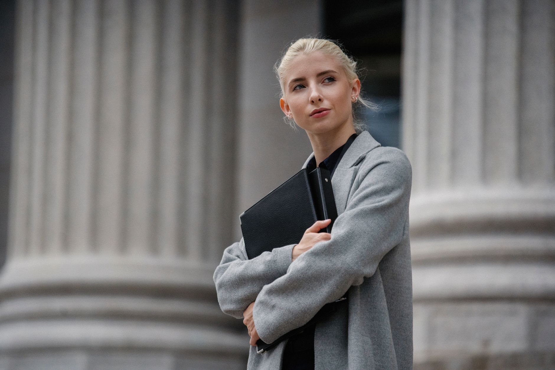 Young self assured blond female executive standing with crossed arms and folders while looking away near stone columns
