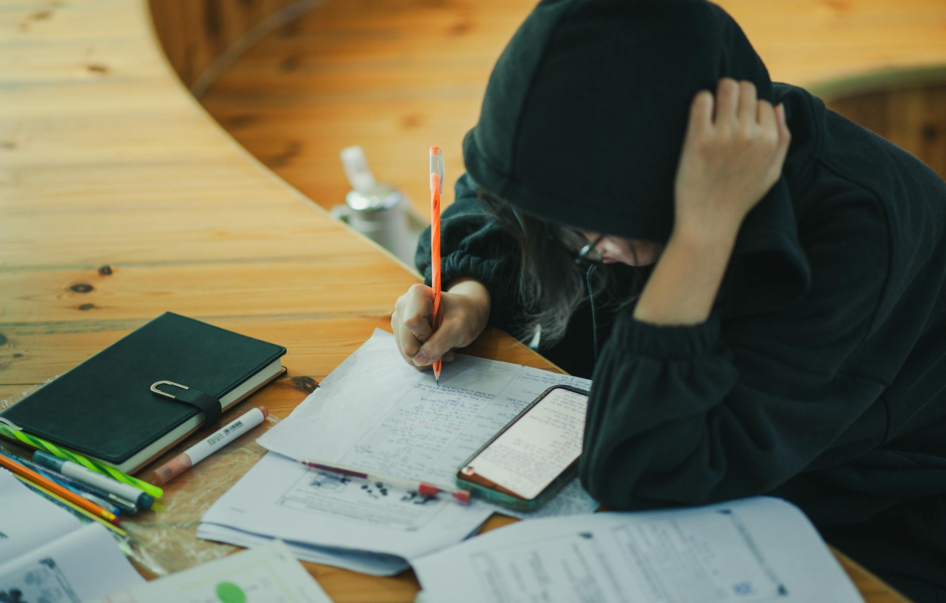 A student wearing a hoodie is deeply engaged in writing notes at a wooden study desk, surrounded by educational materials.