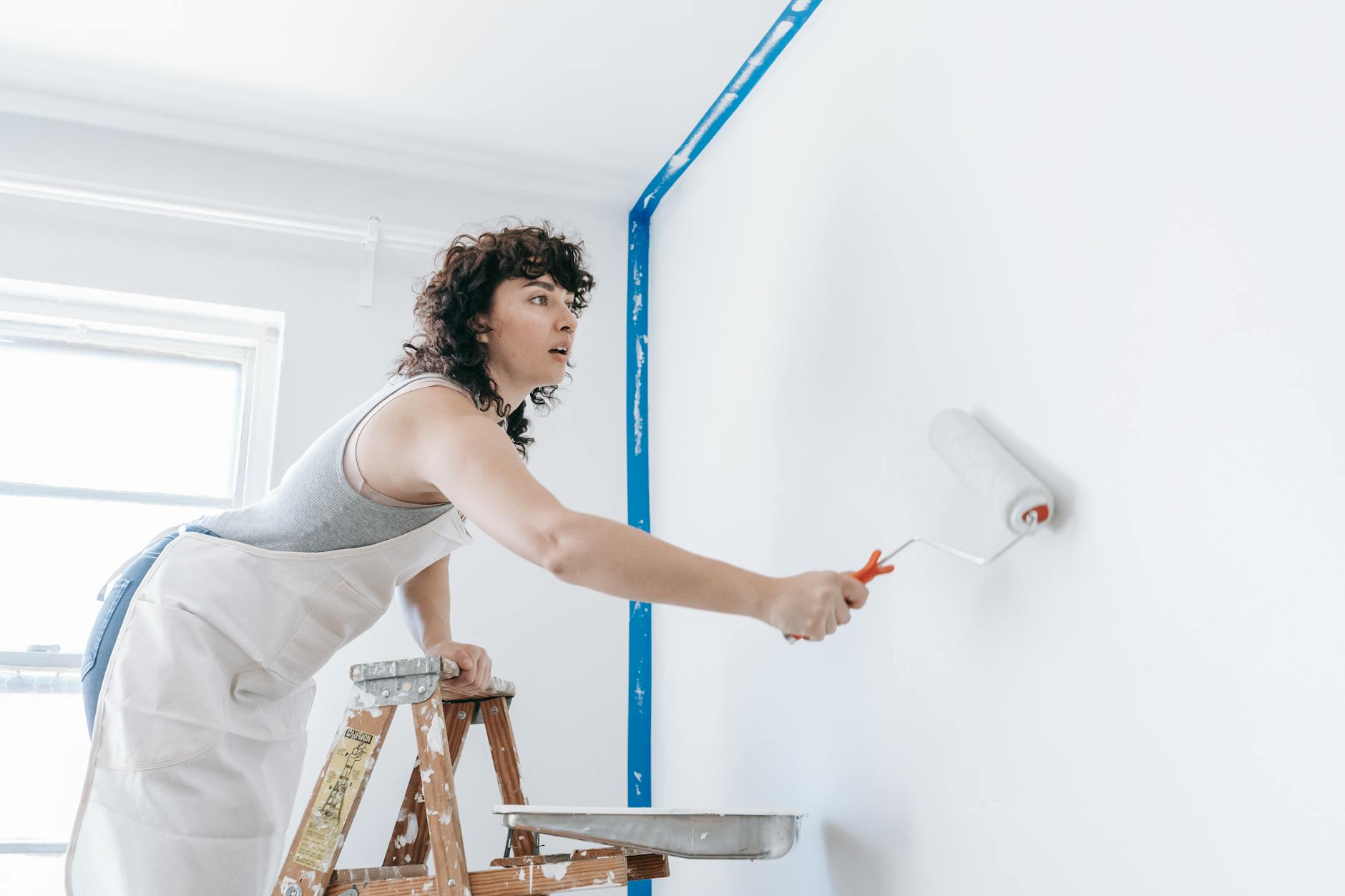 A woman stands on a ladder painting a wall white, part of a DIY home renovation.