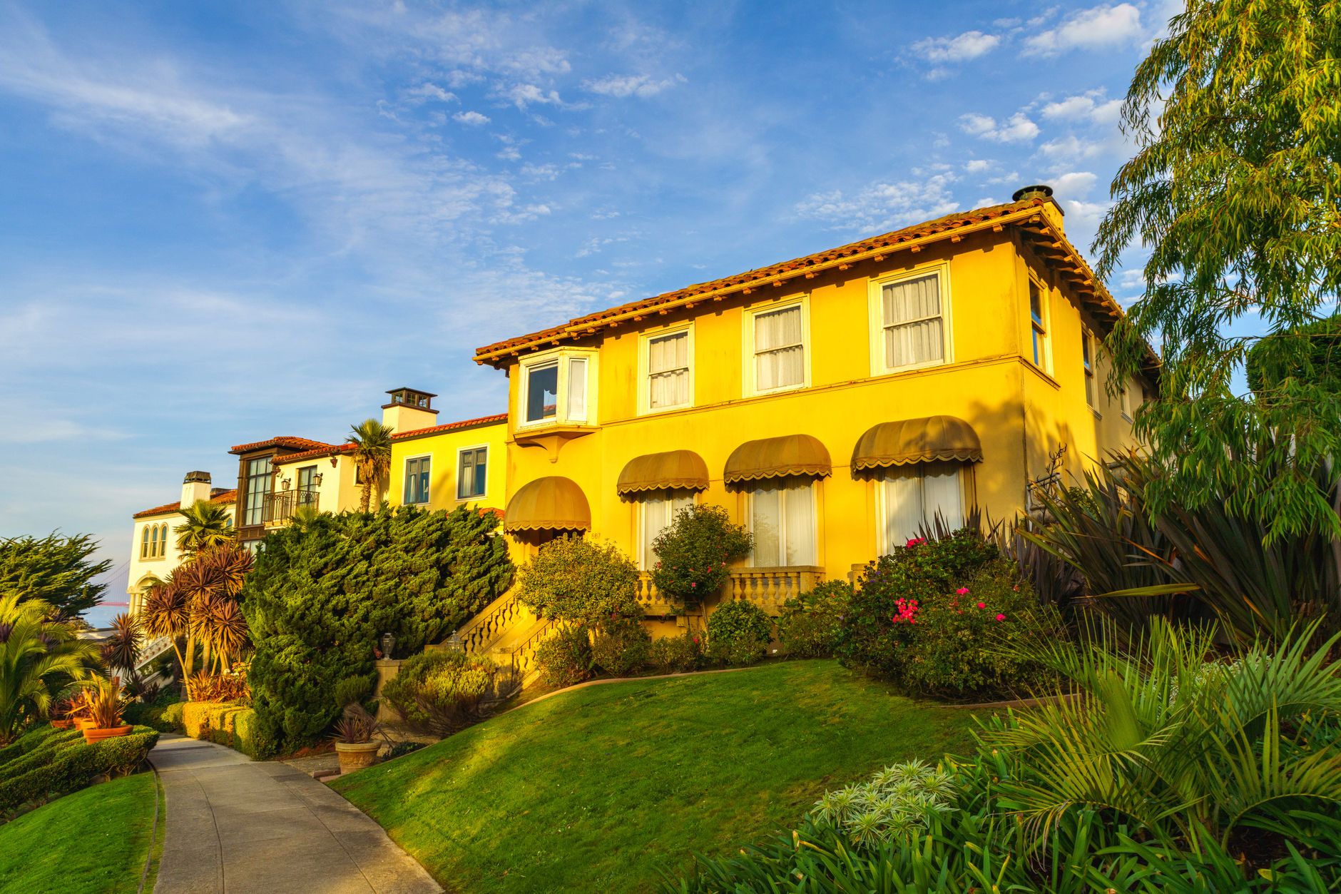 Sunny view of a Mediterranean-style villa surrounded by lush greenery and blue sky.