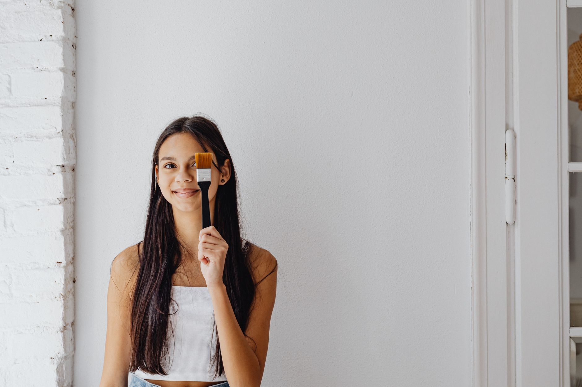 Young woman smiling playfully while holding a paintbrush indoors against a white wall.