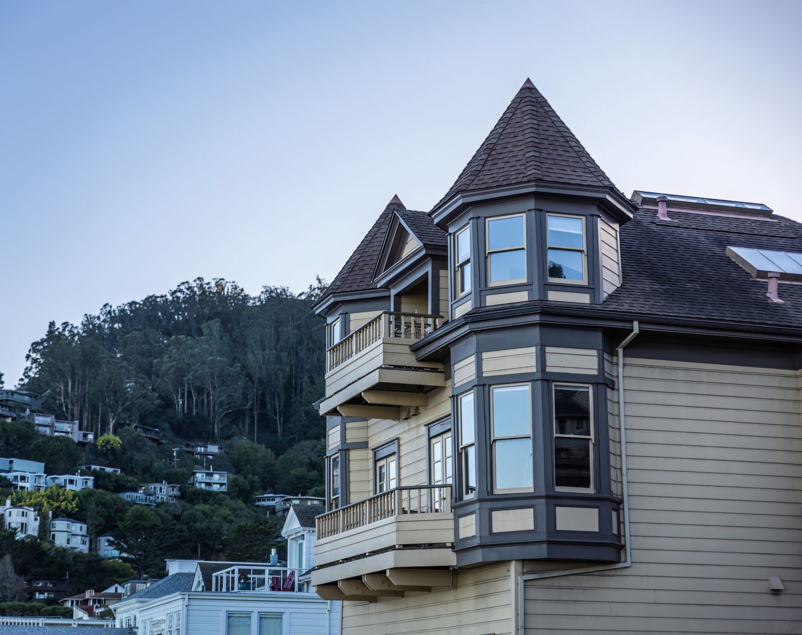 Victorian-style house with wooden balconies against a forested hillside under a clear sky.