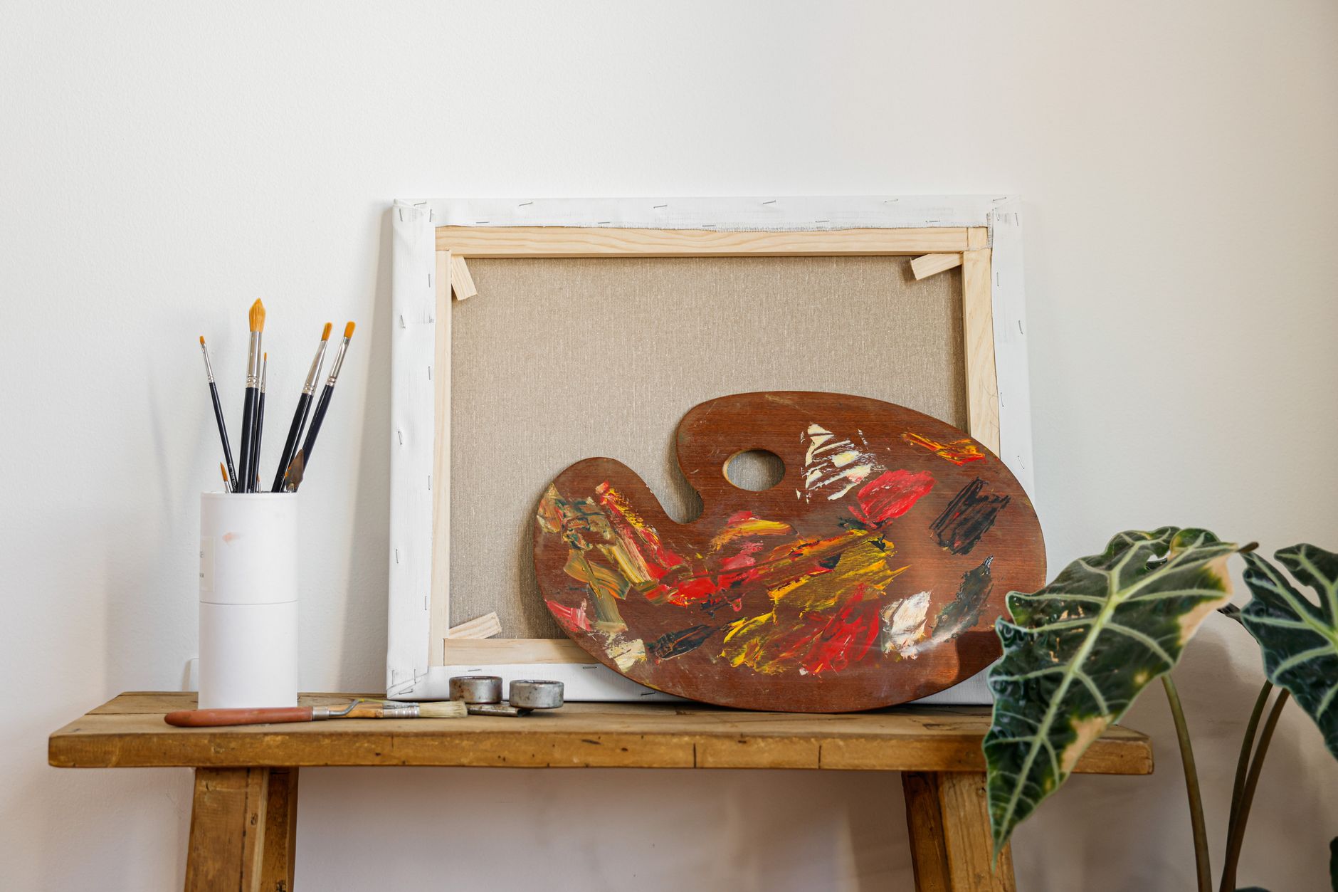 Colorful artist palette and brushes on a wooden bench beside a blank canvas and indoor plant.