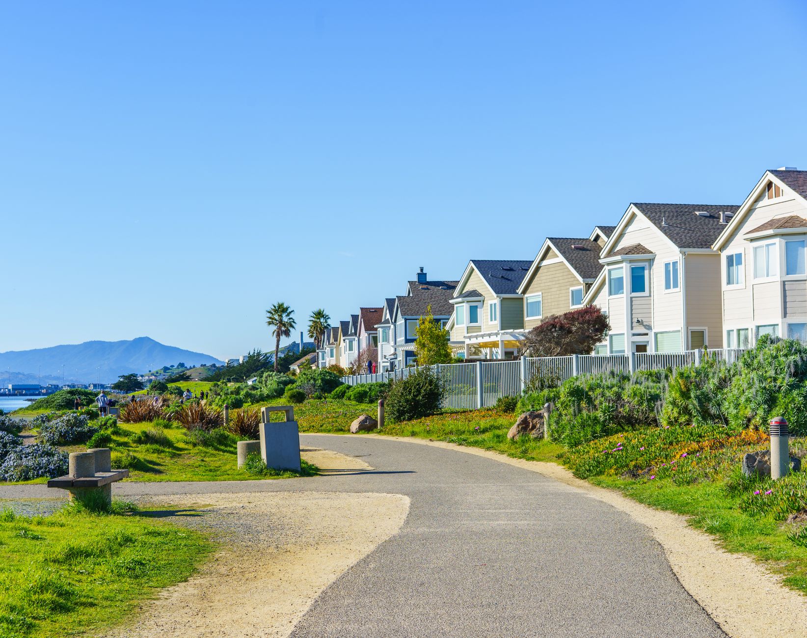 Bright daytime view of a coastal neighborhood with a scenic path and distant mountain backdrop.
