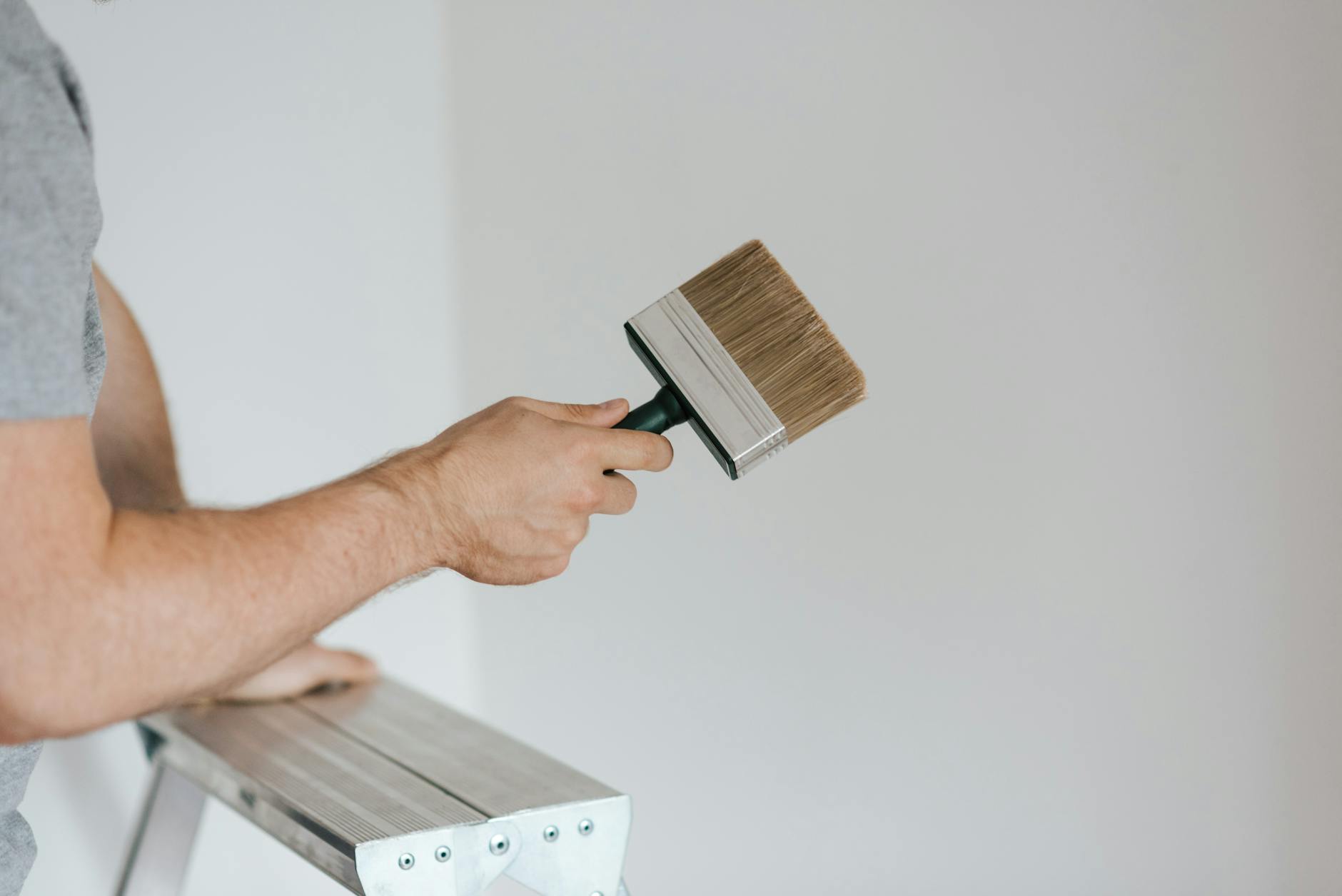 Side view of crop faceless painter in casual clothes holding paintbrush while leaning on stepladder on gray background in soft daylight