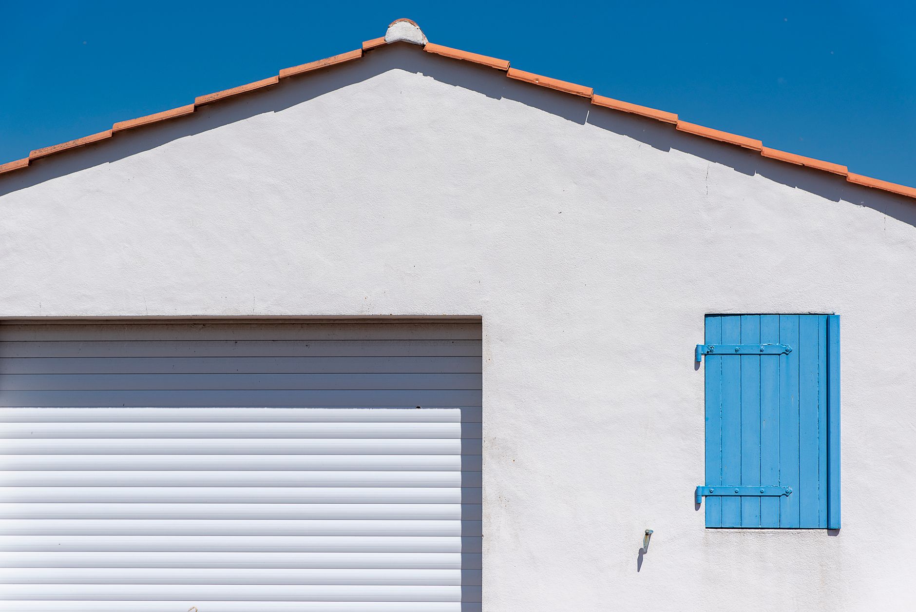 White wall with a blue shutter under a clear blue sky, showing minimalist architecture.