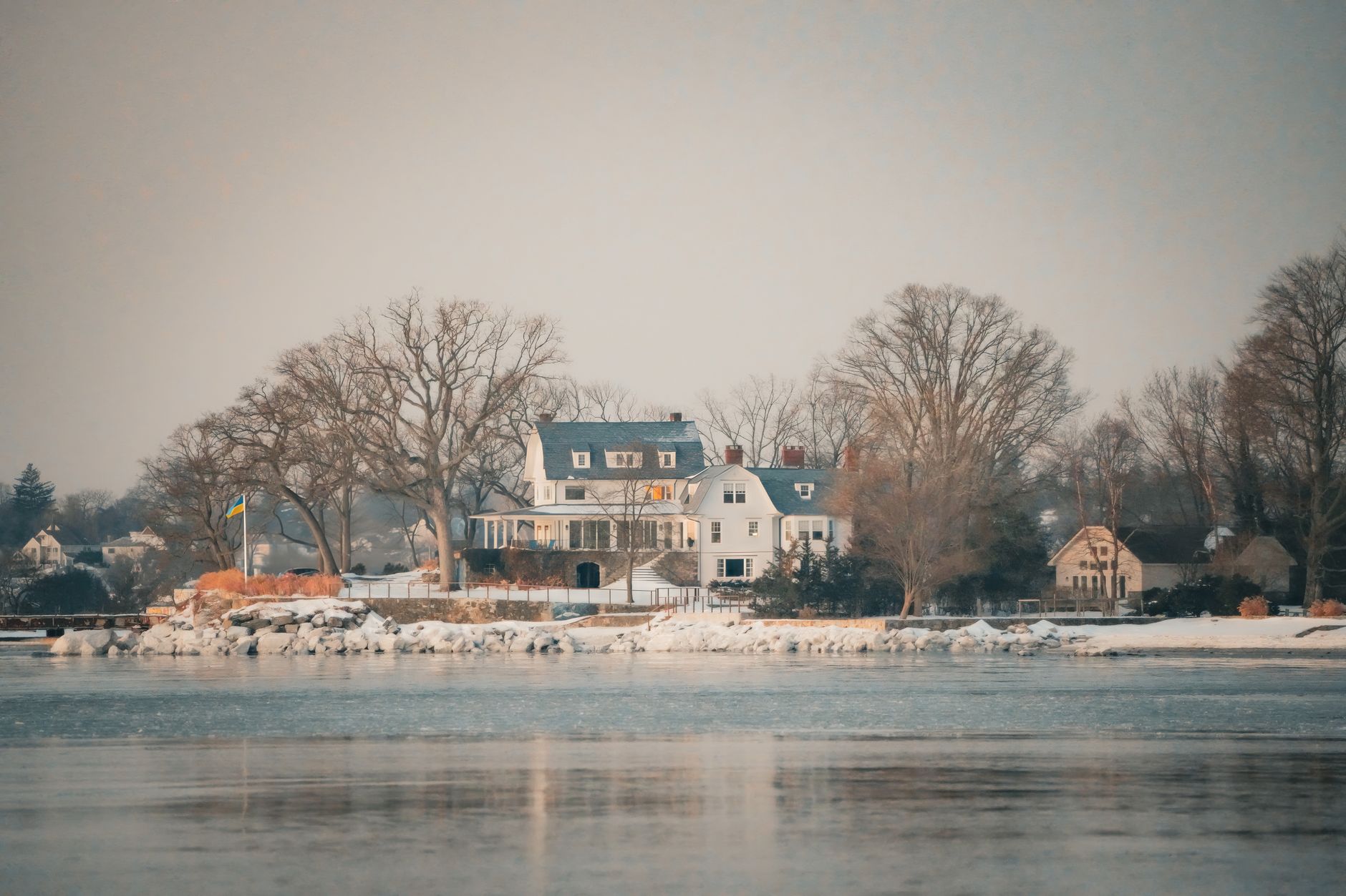 A picturesque lakeside mansion surrounded by snow and bare trees during winter, reflecting calmness.