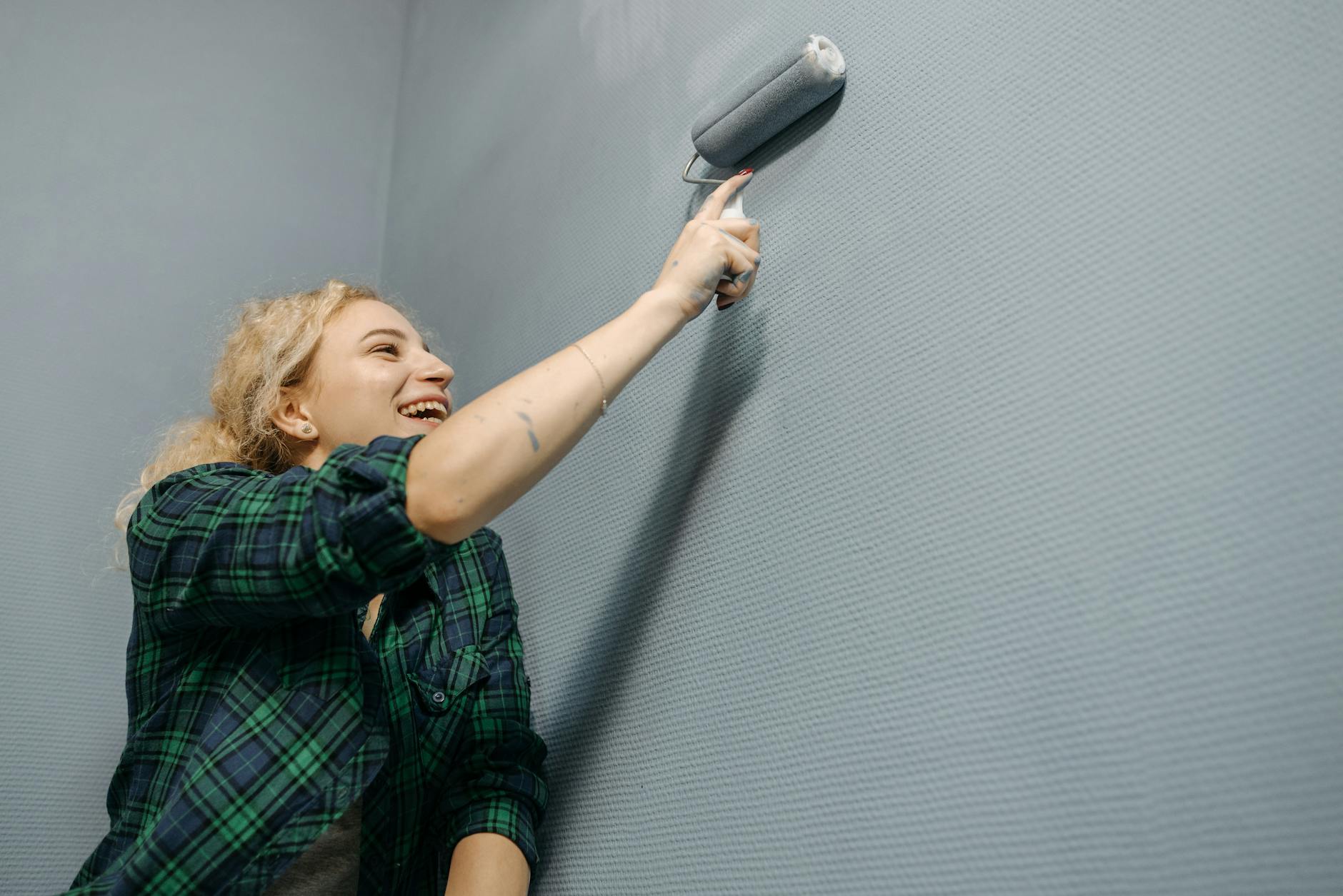 Laughing woman painting a blue wall indoors with a roller brush during home renovation.