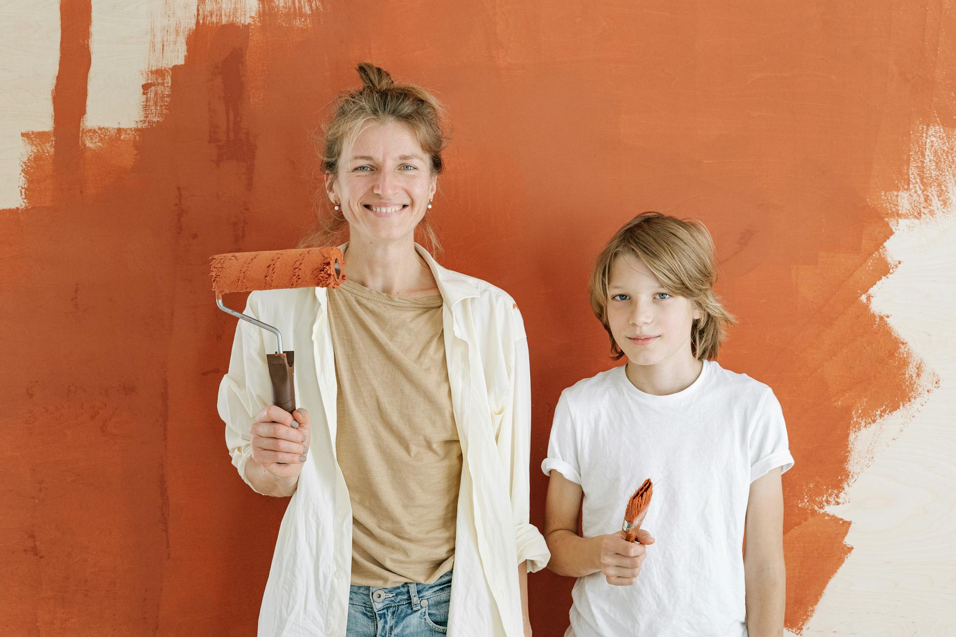 A mother and son smiling while painting a wall indoors, holding paint roller and brush.
