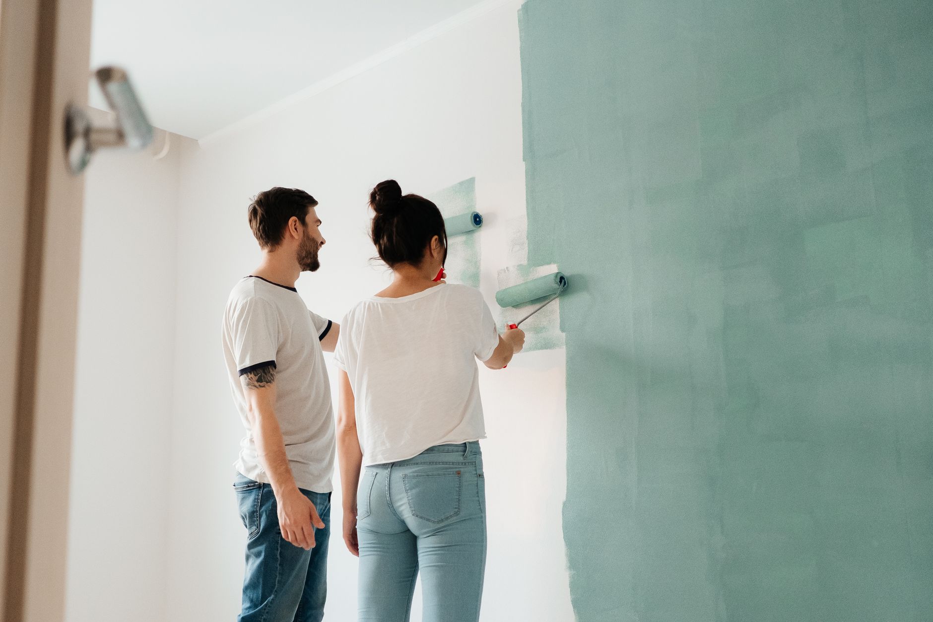 A couple collaborates to paint a wall with a roller indoors, showcasing teamwork.