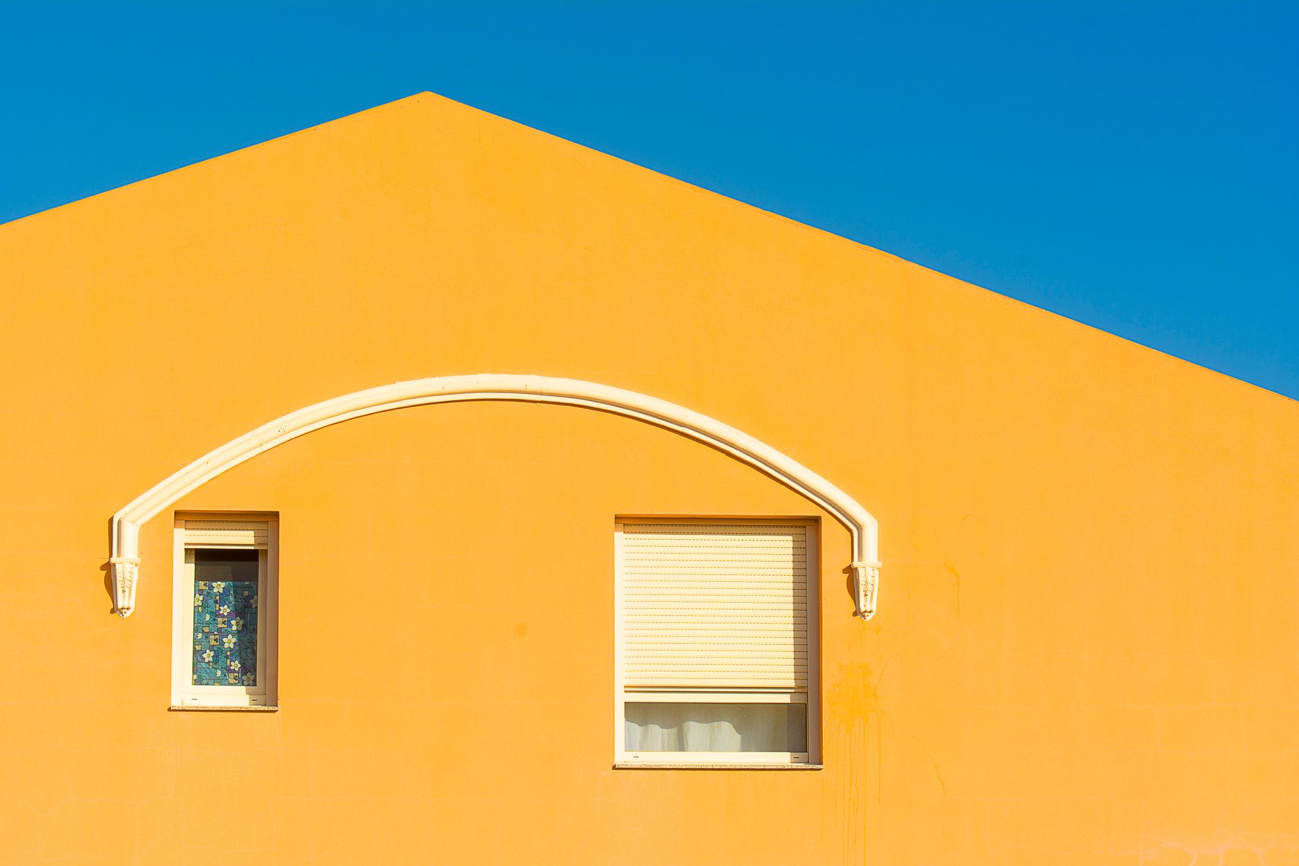 Bright yellow facade of a house with two windows under a clear blue sky, highlighting minimalist architecture.