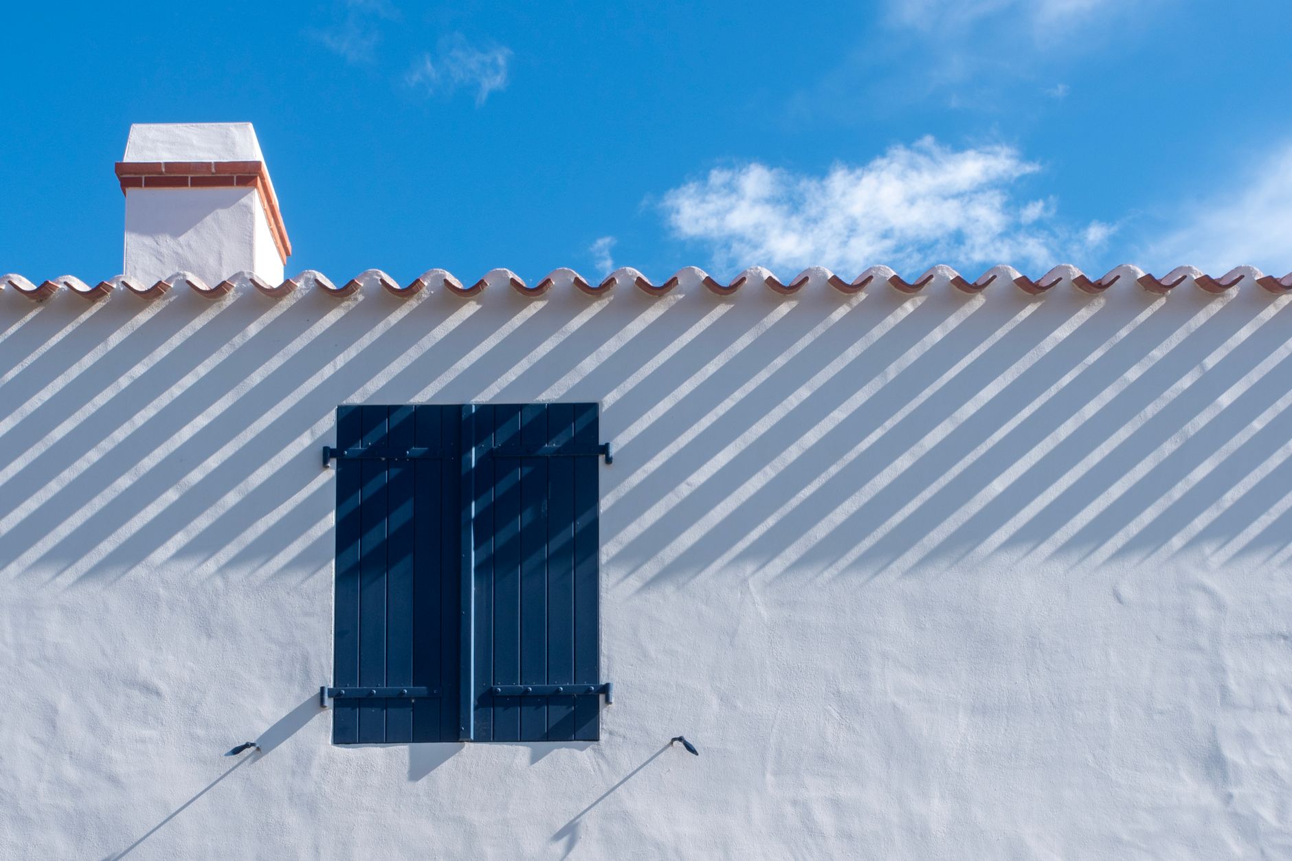 White wall with blue shutters under clear sky casting striped shadows.