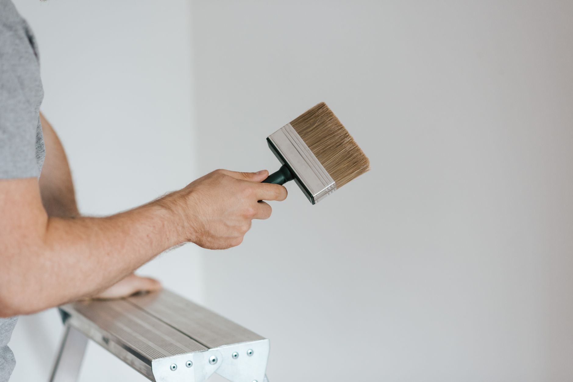 Side view of crop faceless painter in casual clothes holding paintbrush while leaning on stepladder on gray background in soft daylight