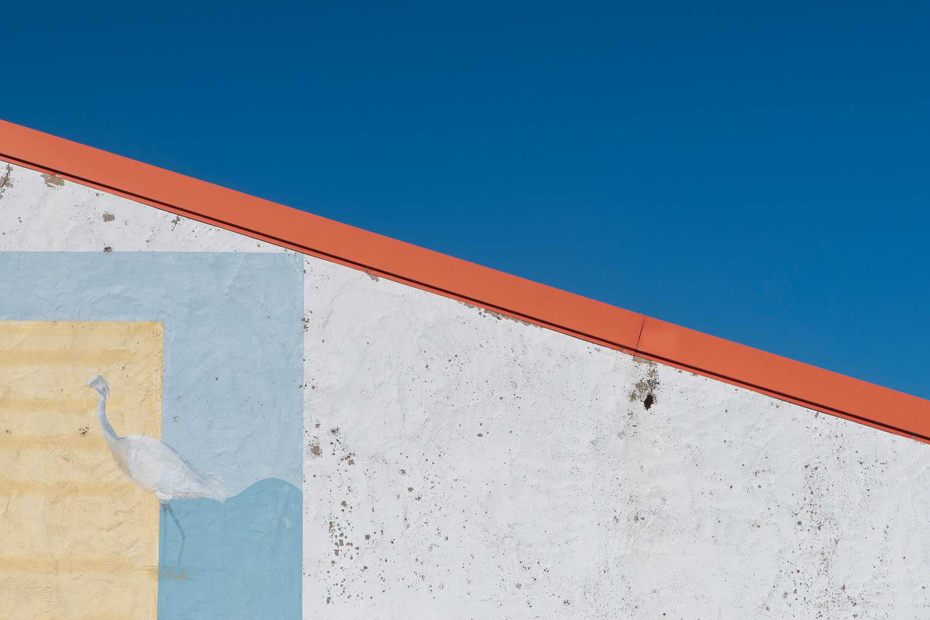 White wall with bird mural contrasts against clear blue sky and diagonal red roofline.