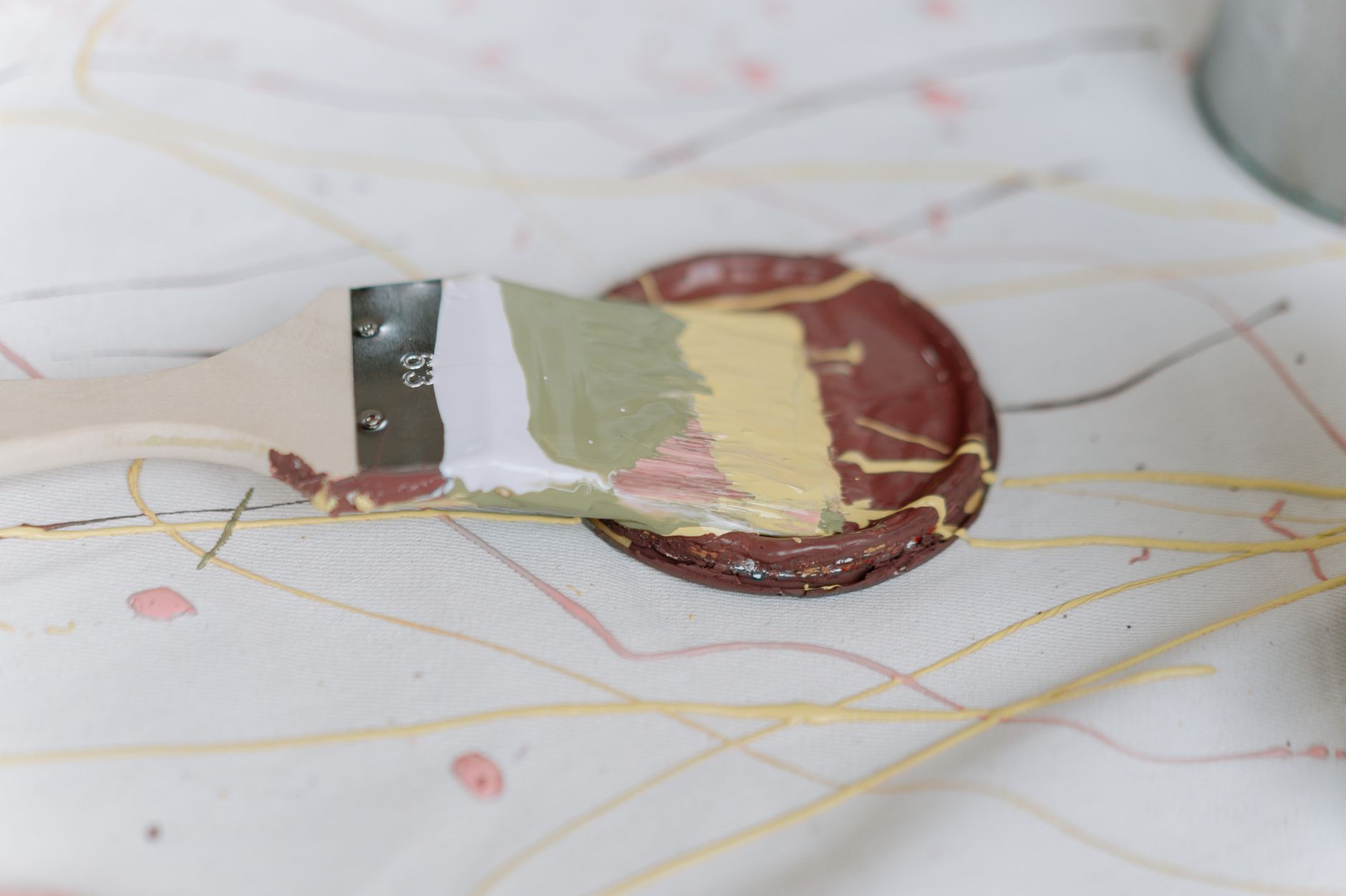 A paintbrush with multiple colors resting on a paint lid atop a decorative canvas.
